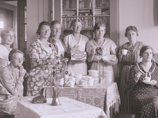 Een groep van acht vrouwen drinkt thee en eet biscuit in een klaslokaal in vermoedelijk Linde.